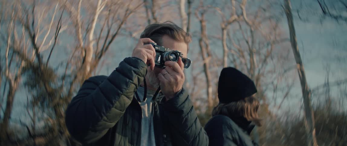 A man with a beard raises a vintage camera to take a photo next to a woman in a wintery forest
