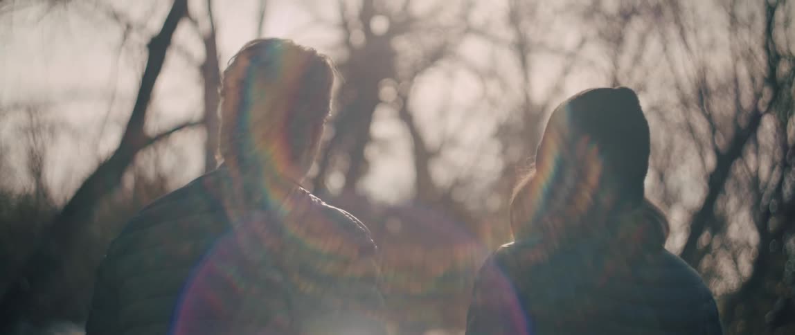 A dreamy, soft-focus shot from behind a couple standing together in a park, backlit by the warm sun which creates a beautiful lens flare