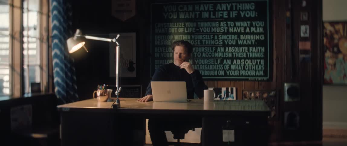 Dolly-in shot of a thoughtful man working on his laptop in a dimly lit, cozy home office with a motivational poster on the wall