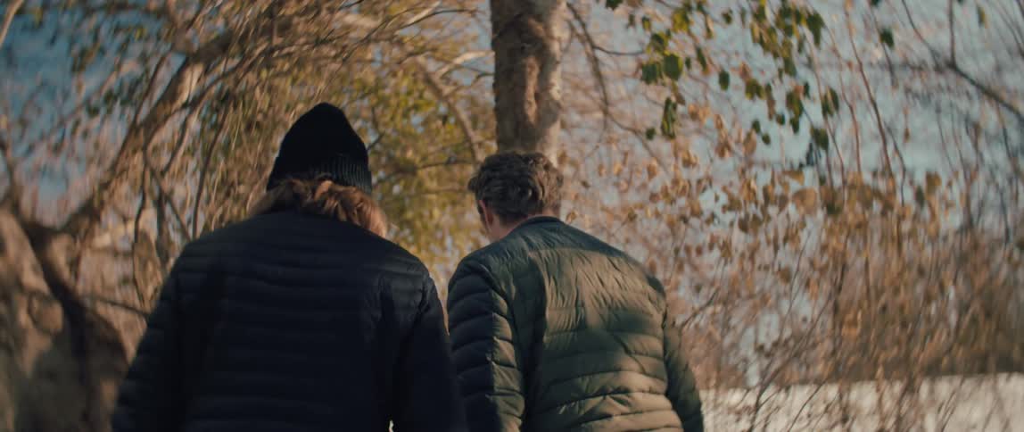 A slow-motion, low-angle shot with a distorted lens follows two men in winter jackets walking through a snowy forest