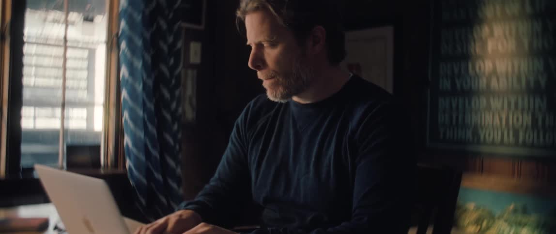 A focused middle-aged man works on his laptop at a desk in a dimly lit room, pausing to put on his glasses to better see the screen