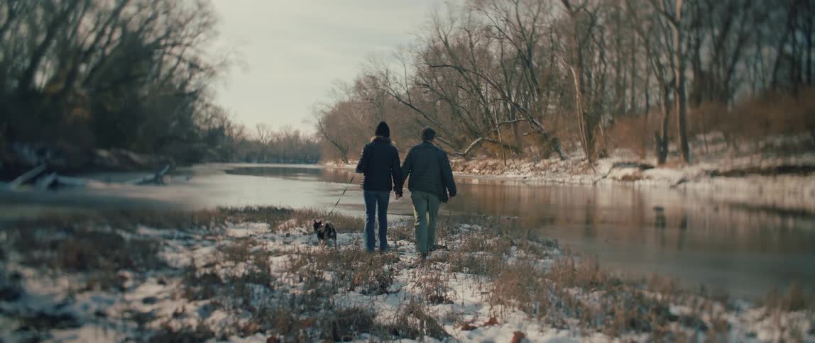From behind, a couple and their dog walk hand-in-hand along a snowy, partially frozen river, stopping to admire the peaceful winter landscape