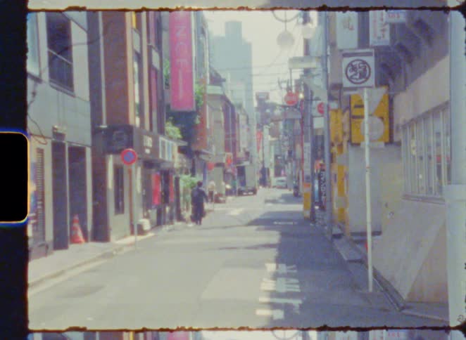 Vintage-style film captures a quiet, sunlit alleyway in a Japanese city with people walking in the distance