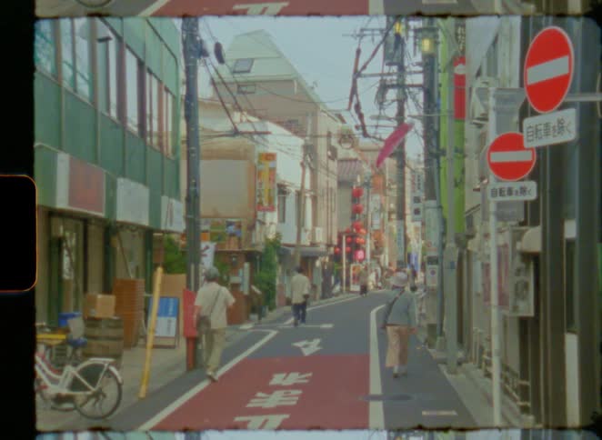People walk and cycle down a narrow street in Japan in this vintage, film-bordered shot that slowly zooms out