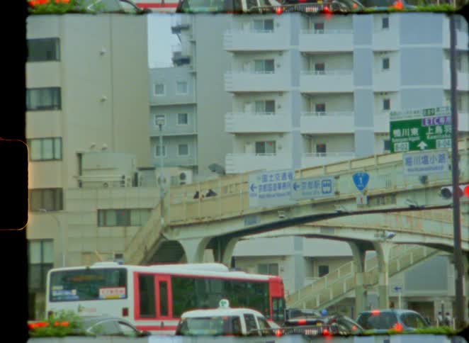 A vintage 8mm film captures a bustling street scene in Japan with a bus passing by and traffic under a footbridge