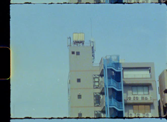Super 8 film scan of a gray apartment building with a bright blue fire escape on a sunny day