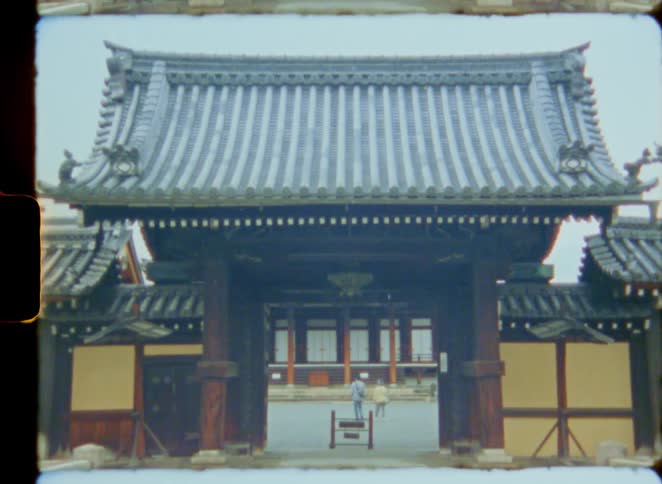 An archival shot on 8mm film zooms out from two people exploring a traditional Japanese temple courtyard to reveal the ornate wooden entrance gate