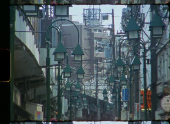 A vintage 8mm film view down a bustling Japanese street lined with unique green lamps as a train passes in the distance