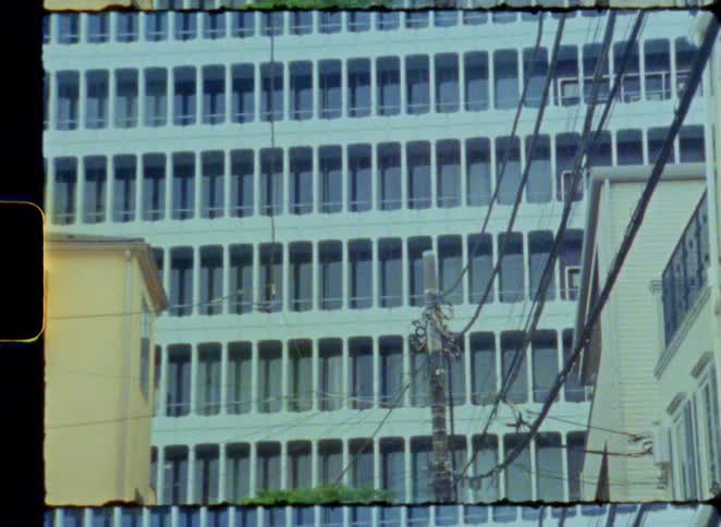 A vintage 8mm film shot zooms out from a telephone pole to reveal a modern office building and a tangle of urban power lines