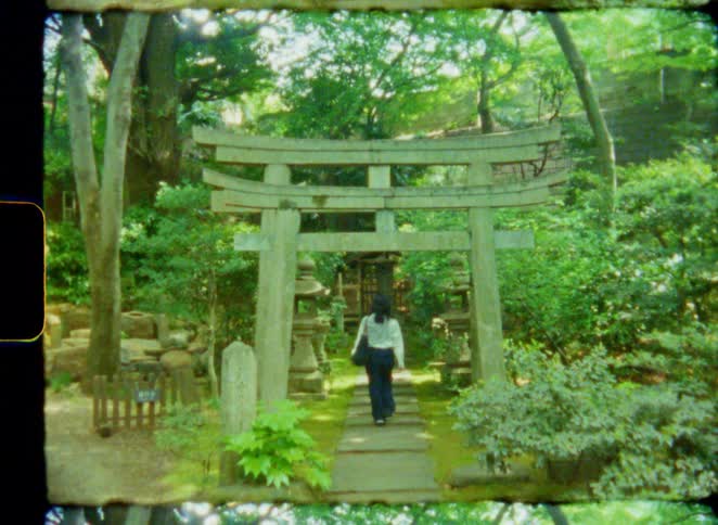 A woman walks through a stone torii gate in a lush Japanese garden towards a shrine in this vintage 8mm style footage