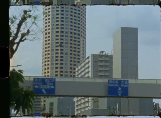 An 8mm vintage film shot slowly zooms in on highway traffic signs against a backdrop of modern Tokyo skyscrapers