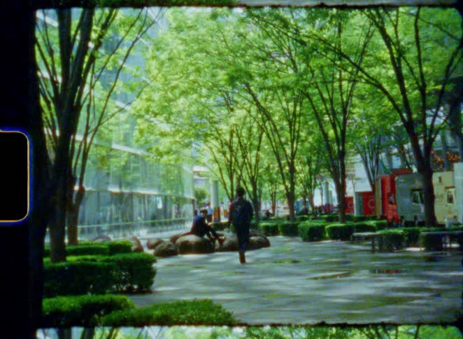 Vintage 8mm footage of men relaxing and walking through a modern urban park with lush green trees and architecture