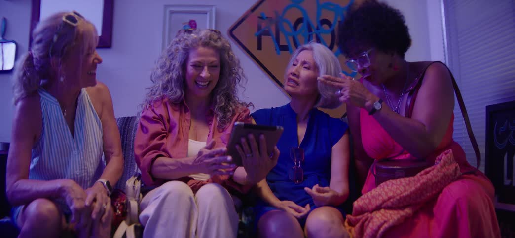 A diverse group of older women friends laugh and joke together while looking at a tablet on a couch