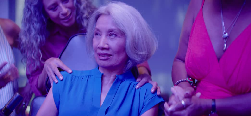 Surrounded by supportive friends, an older woman gets her first tattoo in a stylishly lit parlor