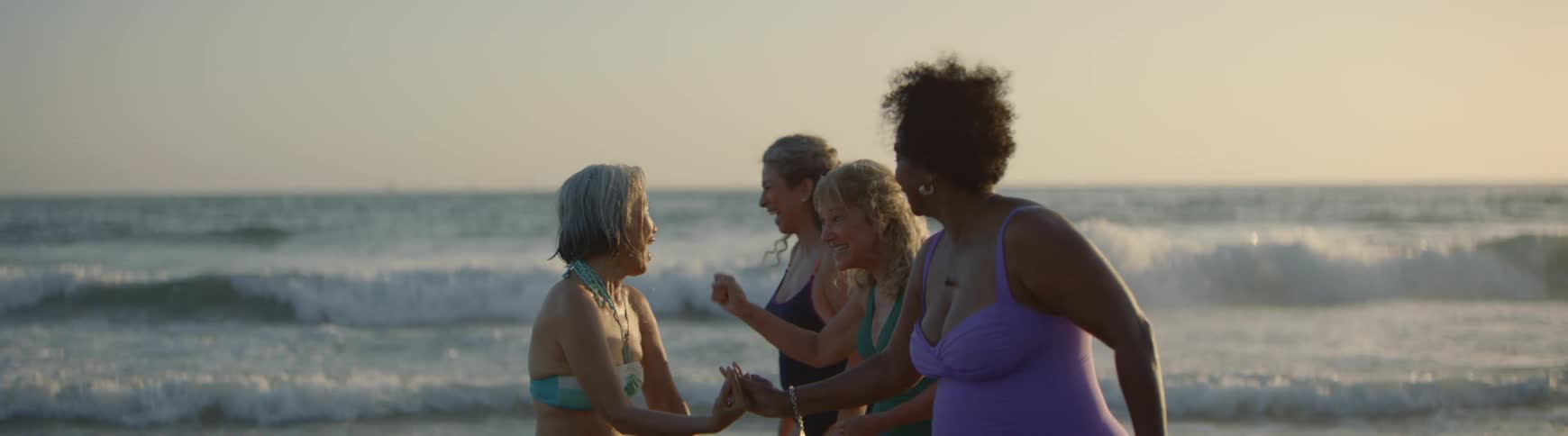 A diverse group of senior women friends in swimsuits laugh and high-five while dancing on the beach at sunset in slow motion