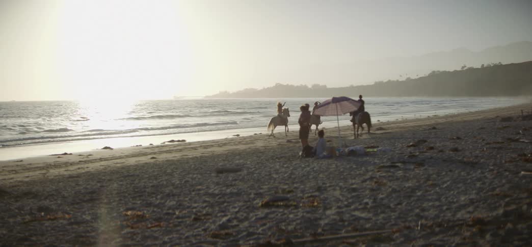 People enjoy a scenic horseback ride along the ocean shore at sunset while a family picnics nearby