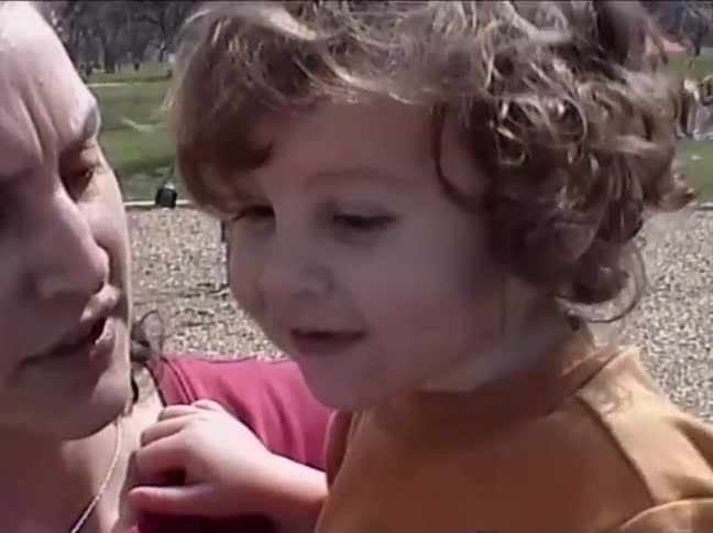 Vintage home video footage of a smiling curly-haired toddler pointing at the camera before sharing a sweet kiss with their mother at the park