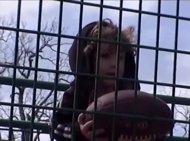 Low angle vintage footage of a young boy in a hoodie holding a football behind a tall green fence at a park