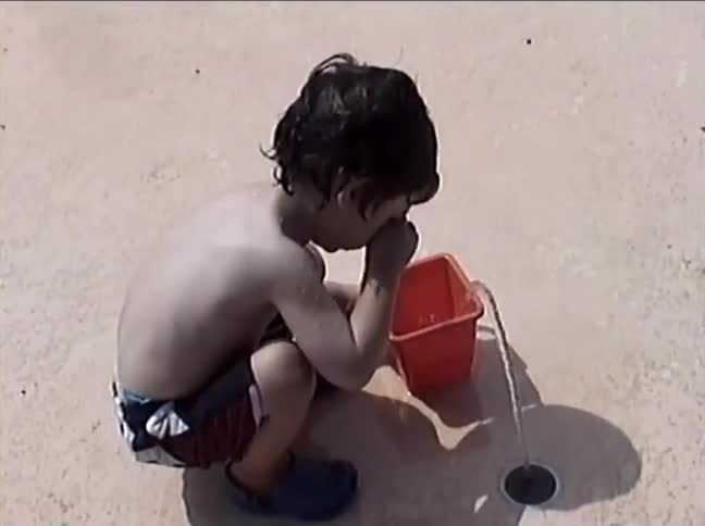 A cute little boy in swim trunks plays with a red bucket at a splash pad, filling it with water from a ground fountain and smiling at the camera