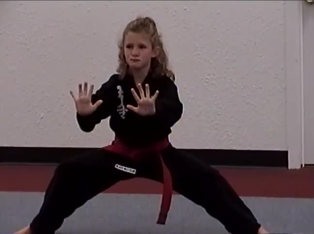 A focused young girl in a black gi and red belt holds a martial arts stance in a dojo