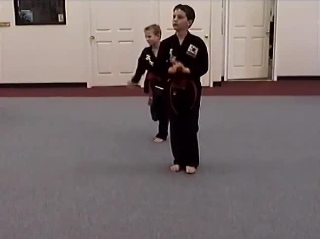A focused young boy in a black uniform with a red belt practices a choreographed martial arts routine in a dojo