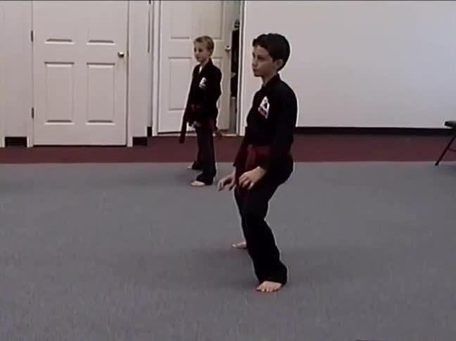 A group of young children in black uniforms with red belts practice a synchronized martial arts kata in a dojo