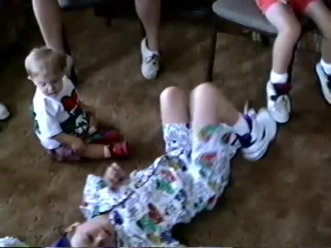 A nostalgic home video captures a young boy in a blue hat doing sit-ups on the carpeted floor while a toddler watches