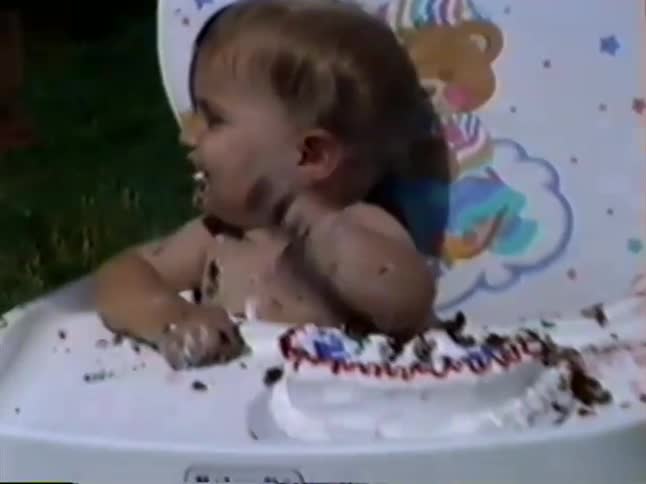 A joyful toddler covered in frosting laughs and plays with her smash cake during an outdoor birthday party