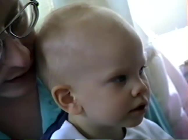 Vintage home video footage zooms in on a serious-looking baby boy with blue eyes being held by his parents on a couch