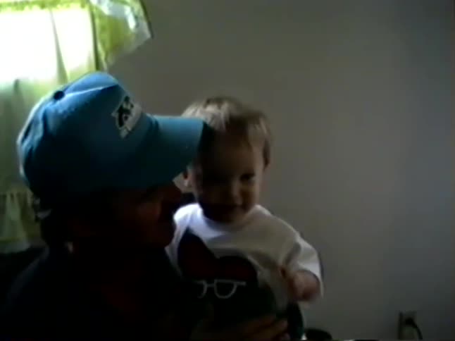 A nostalgic home video captures a tender moment as a father holds, kisses, and changes his young son's shirt indoors