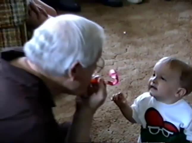 A cute baby laughs and plays with their grandmother and a party horn blower toy in a nostalgic home video