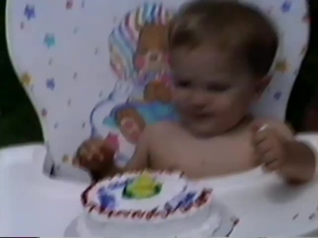 A toddler sitting in a high chair screams during their birthday celebration before calming down to eat their cake