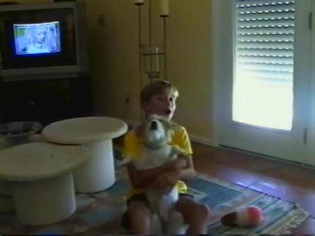 Nostalgic home video footage shows a young girl in a yellow t-shirt lovingly holding her small scruffy dog in the living room