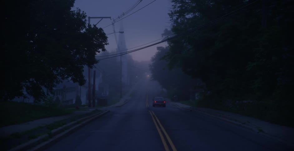 A car drives down a foggy road near dusk