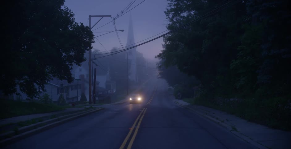 A car drives along a two-lane road in a village at dawn