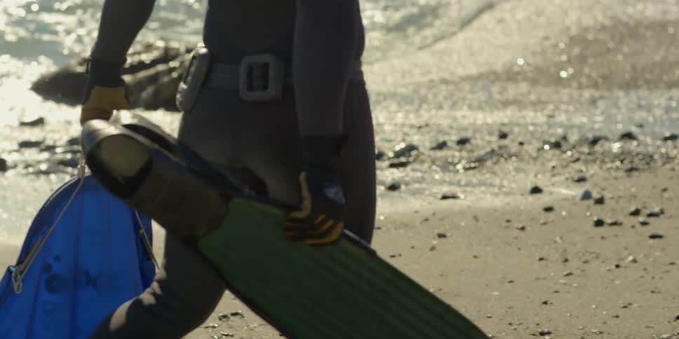 A man in a wetsuit walks on the beach carrying flippers and a net