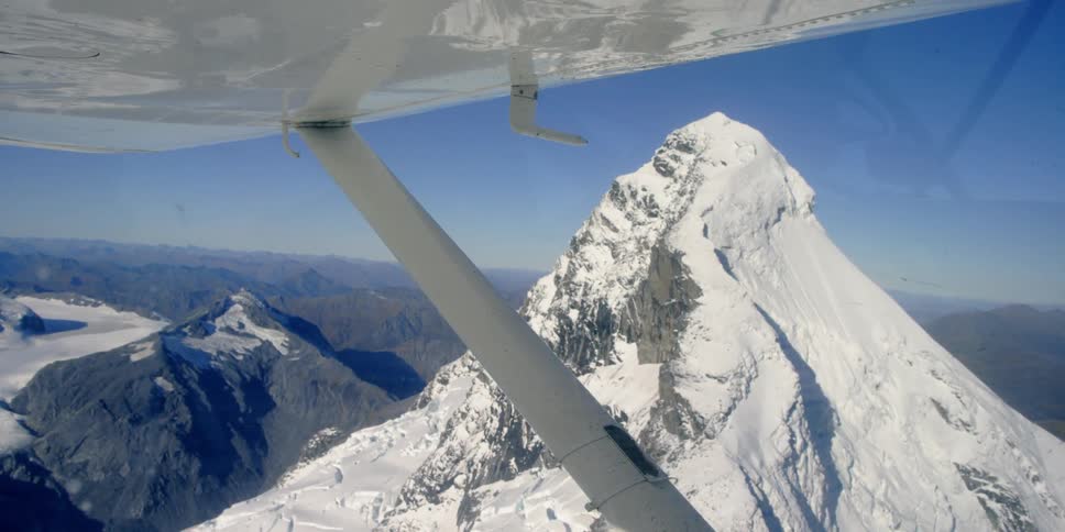 A plane flies above a tall mountain peak on a sunny day