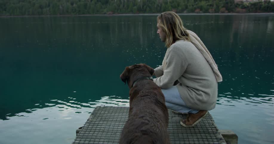 A man and a dog rest on the end of a dock at a lake
