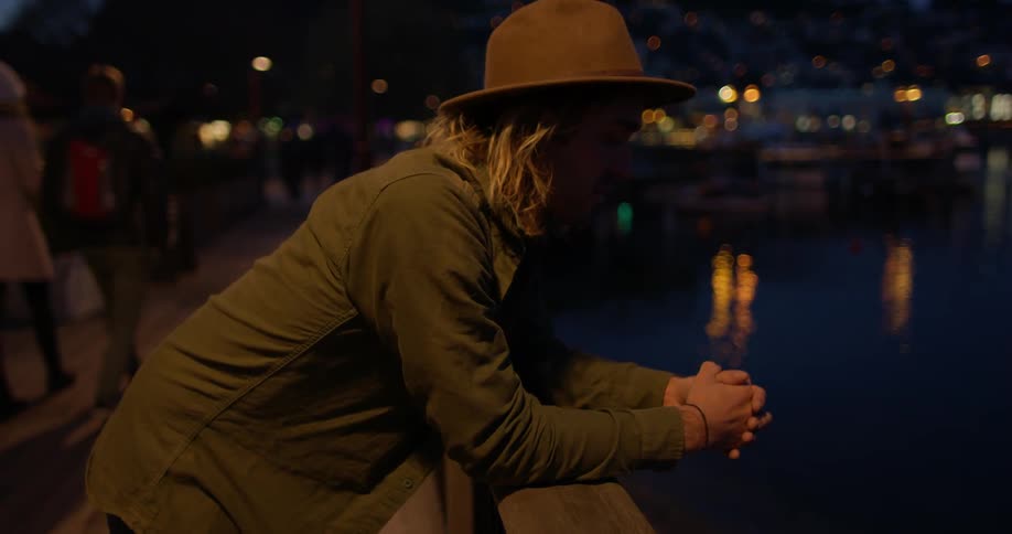 Man leaning on a rail next to water wearing a hat at dusk