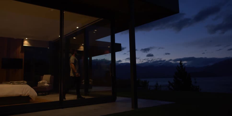 A man looks out the window of a lake house at dusk