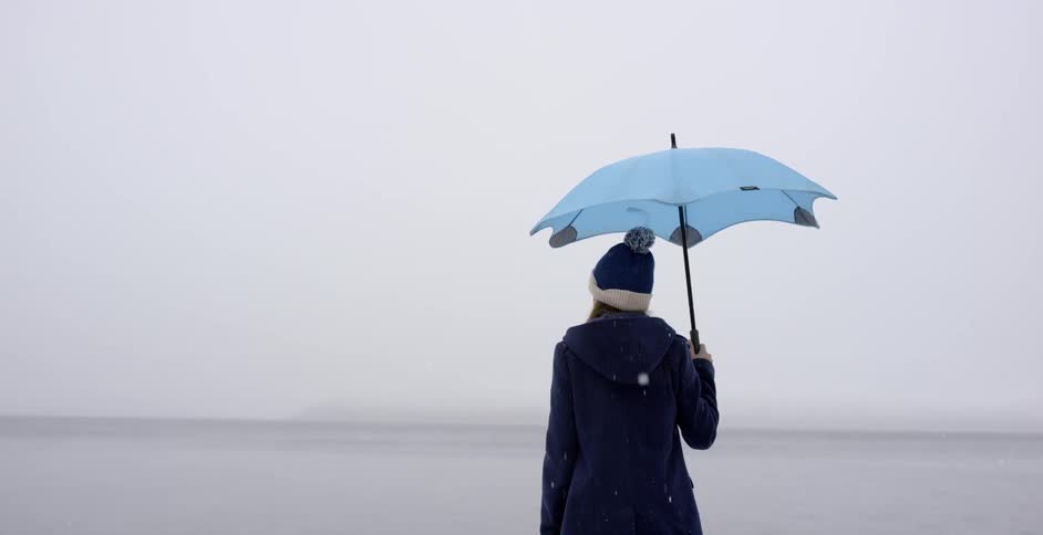 A person holds a blue umbrella by the water as it snows