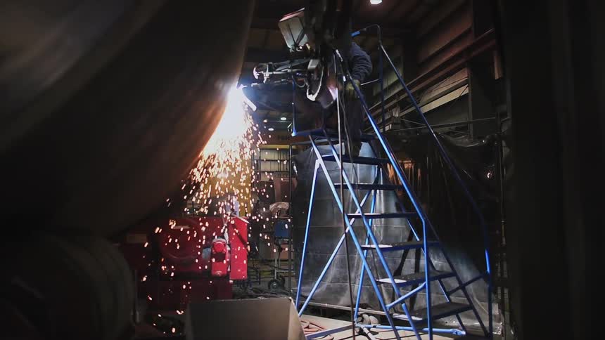 A worker uses a power tool on a large metal cylinder as sparks fly