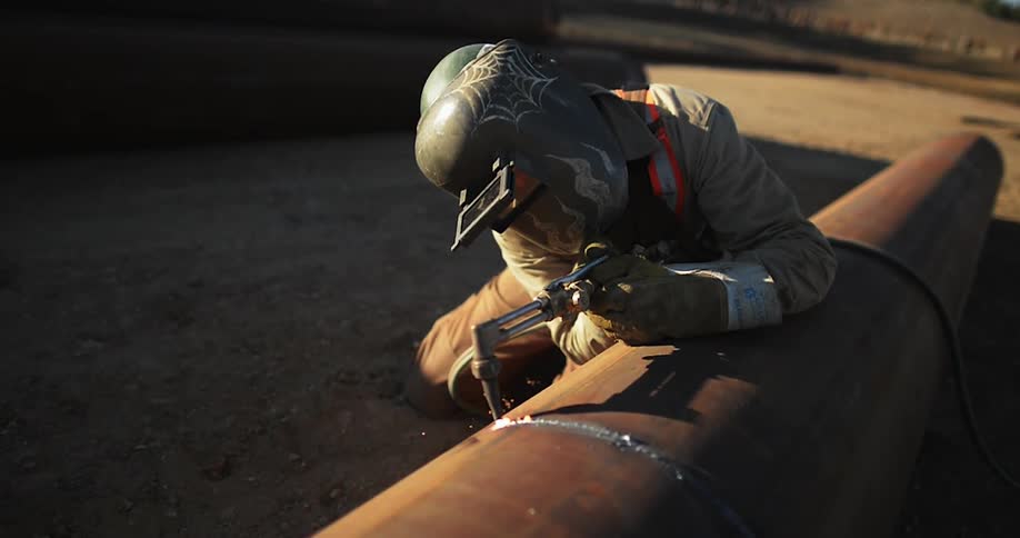 A worker cuts a large metal pipe while sparks fly