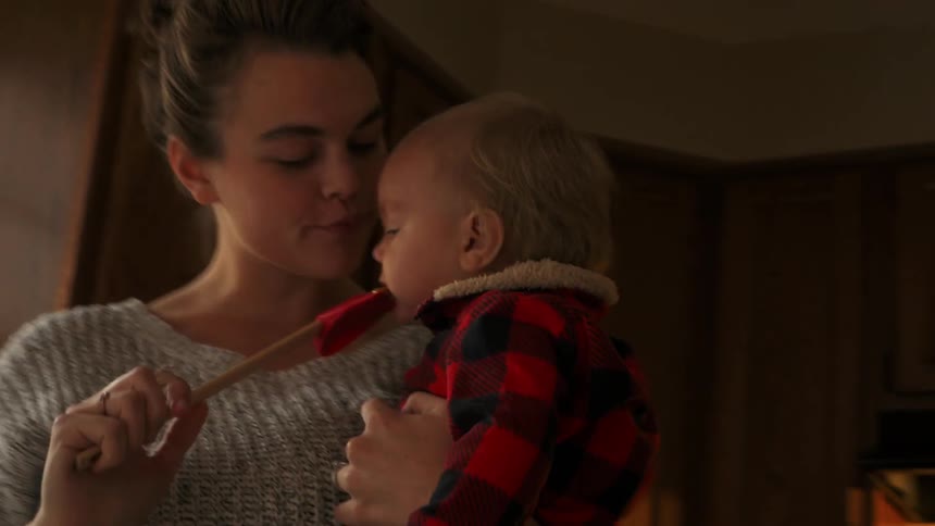 Woman carries a baby in a kitchen and licks spatula