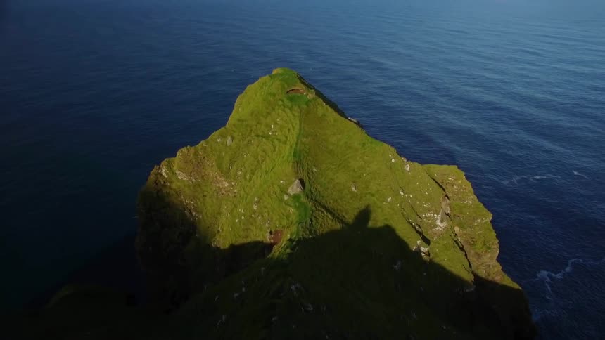 A small tower is on the height of a green mountain by the water