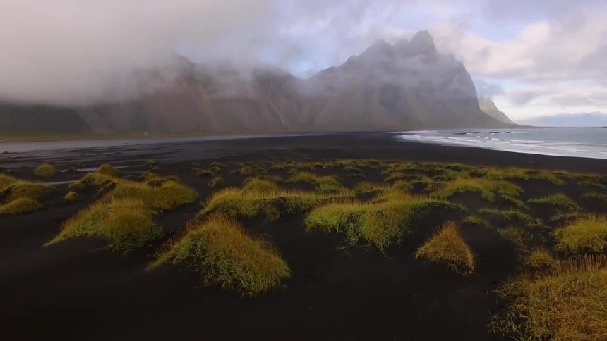 flight over grass topped black sand dunes with ocean and mountain range in background