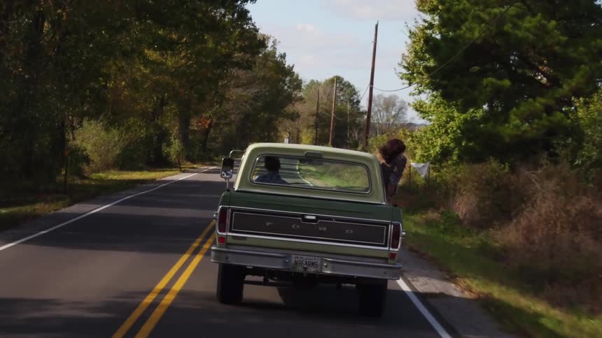 A woman leans out the window of a moving pickup truck in a rural area