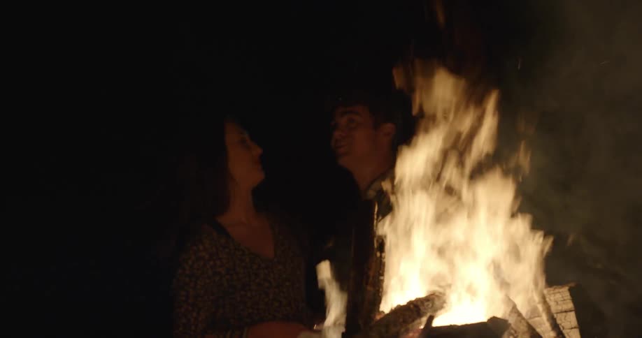 Man and woman sit in front of a fire smiling at night