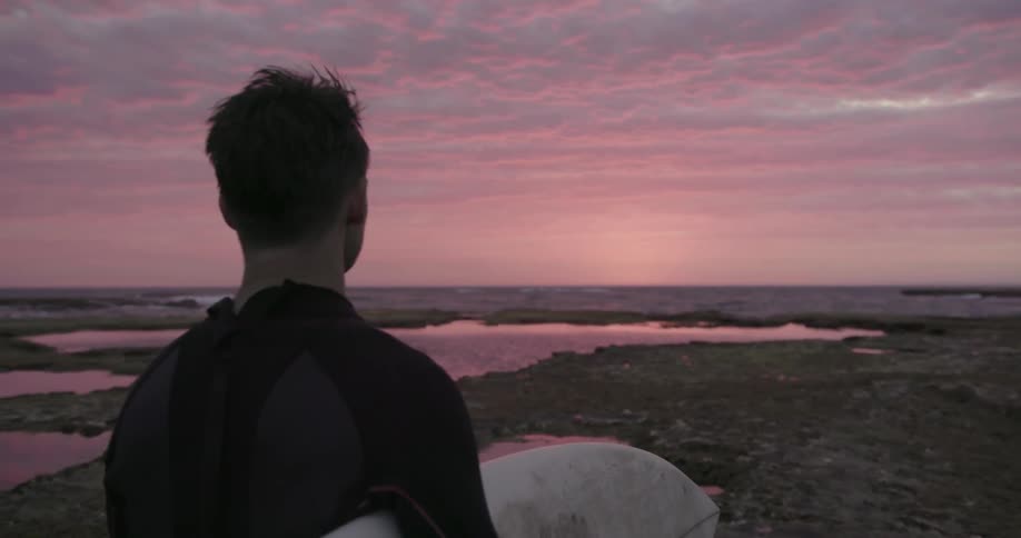 A surfer stands on a beach at sunset