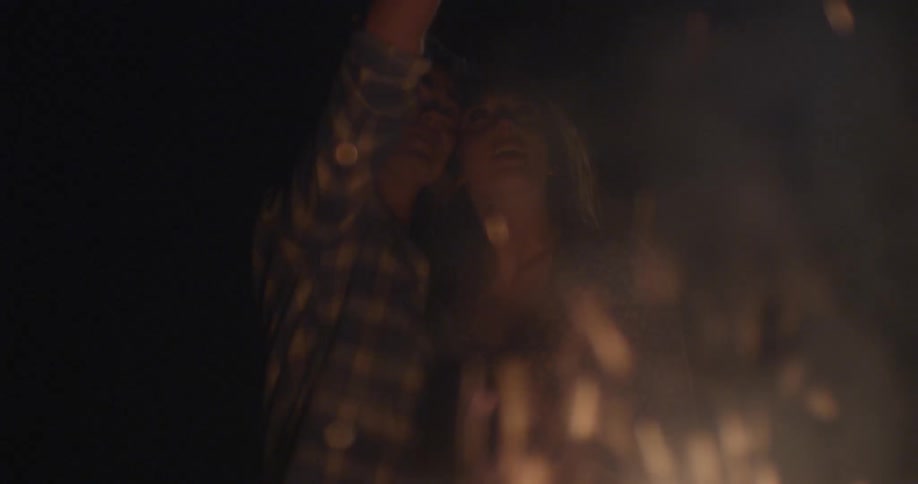 Man and woman sit in front of a fire smiling at night